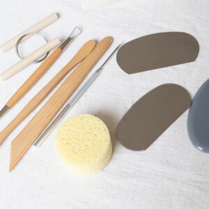 May include: A set of pottery tools including a wooden modeling tool, a metal sculpting tool, a sponge, and a metal scraper. The tools are laid out on a white fabric surface.
