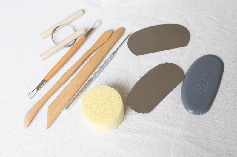 May include: A set of pottery tools including a wooden modeling tool, a metal sculpting tool, a sponge, and a metal scraper. The tools are laid out on a white fabric surface.