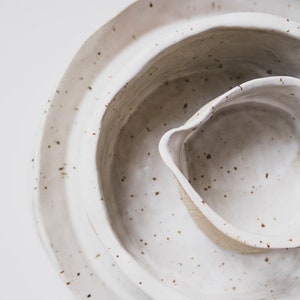 Op de afbeelding: Een set van drie witte keramische kommen met gespikkelde bruine glazuur. De kommen zijn in elkaar genest, met de kleinste kom in het midden.