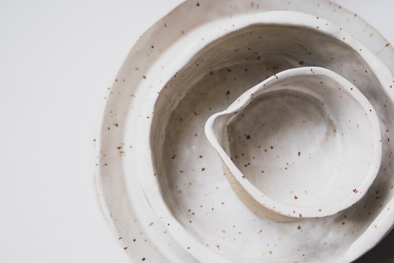 Op de afbeelding: Een set van drie witte keramische kommen met gespikkelde bruine glazuur. De kommen zijn in elkaar genest, met de kleinste kom in het midden.