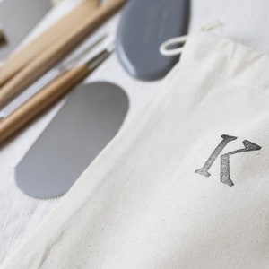 Op de afbeelding: Een set pottenbakkersgereedschap in een witte canvas tas met de letter "K" erop gedrukt. De gereedschappen omvatten een houten handvat, een metalen schraper en een metalen naaldgereedschap.