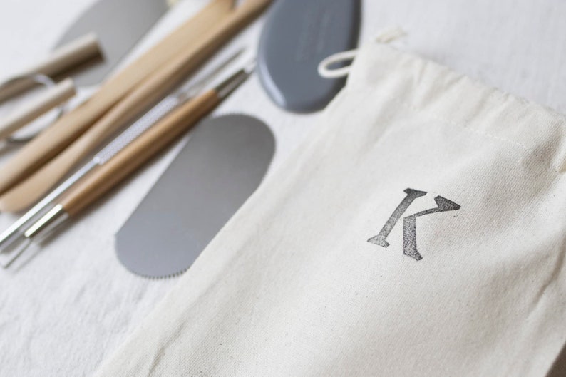 May include: A set of pottery tools in a white canvas bag with the letter "K" printed on it. The tools include a wooden handle, a metal scraper, and a metal needle tool.