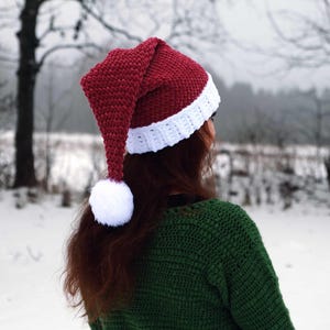 Op de afbeelding: Een rood-witte gebreide kerstmanmuts met een witte pompon aan het uiteinde van de muts. De muts wordt gedragen door een persoon met lang bruin haar. De persoon staat in een besneeuwd landschap.
