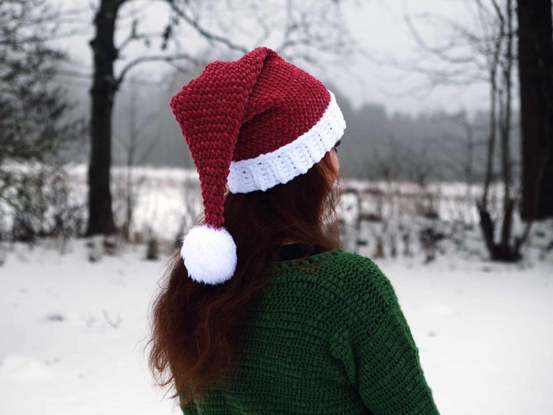 Op de afbeelding: Een rood-witte gebreide kerstmanmuts met een witte pompon aan het uiteinde van de muts. De muts wordt gedragen door een persoon met lang bruin haar. De persoon staat in een besneeuwd landschap.