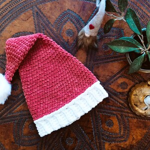 Op de afbeelding: Een roze en witte gehaakte kerstmanmuts met een witte pompon bovenop. De muts ligt op een bruin houten oppervlak met ingewikkelde houtsnijwerk.