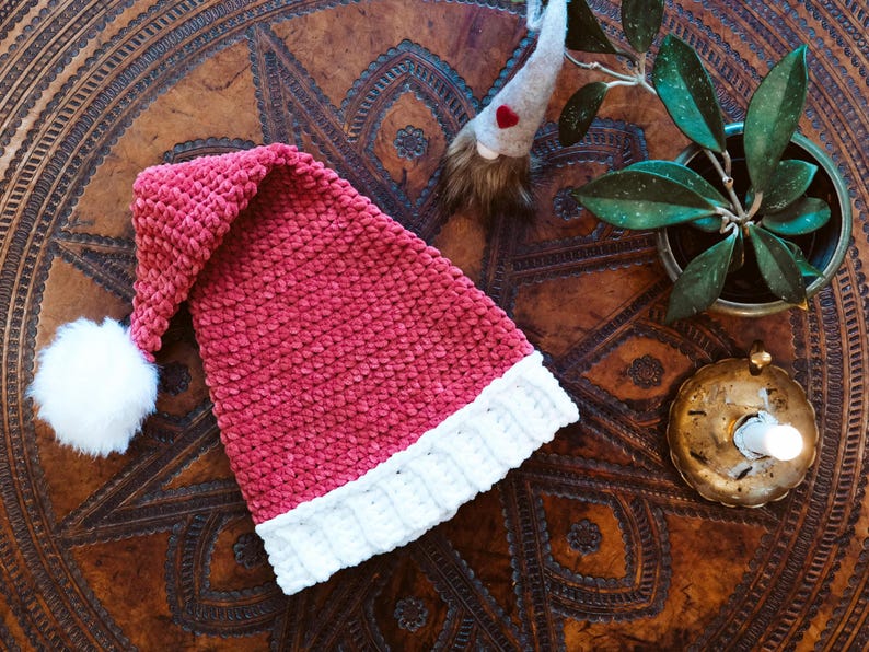 Op de afbeelding: Een roze en witte gehaakte kerstmanmuts met een witte pompon bovenop. De muts ligt op een bruin houten oppervlak met ingewikkelde houtsnijwerk.