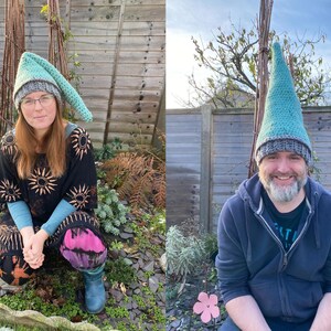 Op de afbeelding: Twee mensen dragen groene en grijze gebreide mutsen in gnome-stijl. De mutsen hebben lange, puntige toppen. De mensen lachen en staan voor een houten hek.