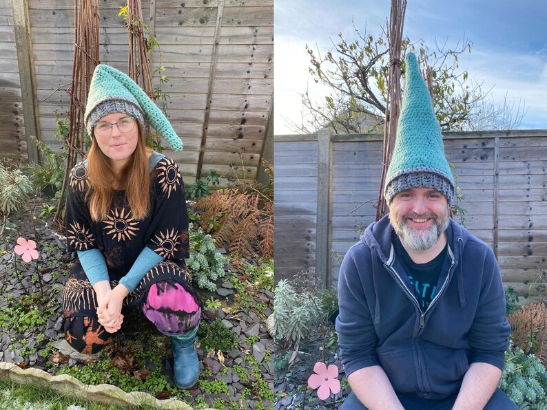 Op de afbeelding: Twee mensen dragen groene en grijze gebreide mutsen in gnome-stijl. De mutsen hebben lange, puntige toppen. De mensen lachen en staan voor een houten hek.