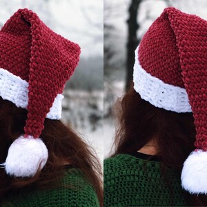 Op de afbeelding: Een rood-witte gebreide kerstmanmuts met een lange, puntige punt en een witte pompon aan het einde. De muts wordt gedragen door een persoon met lang bruin haar.