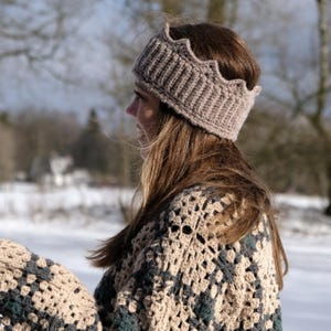 May include: A taupe-colored, crocheted headband with a scalloped edge. The headband is worn by a person with long brown hair. The person is also wearing a crocheted shawl in shades of beige, green, and brown. The background is a snowy, outdoor scene.