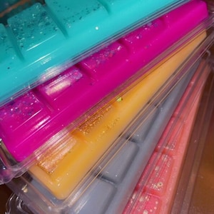 May include: A stack of six wax melts in clear plastic packaging. The wax melts are in various colors, including teal, pink, orange, gray, and pink with white flecks.