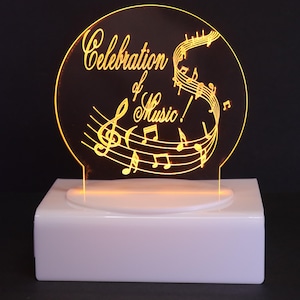 May include: A lighted musical decoration featuring a circular design with the words "Celebration of Music!" in a flowing script. The design includes musical notes and a treble clef, illuminated with a warm yellow glow, set on a white rectangular base.