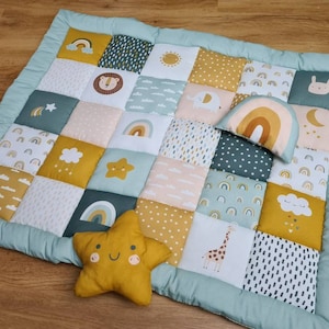 Könnte beinhalten: Eine Patchwork-Spielmatte für Babys mit hellgrünem Rand. Die Quadrate zeigen verschiedene Designs, darunter Regenbogen, Wolken, Sterne und Tiere in Gelb-, Grün- und Rosatönen. Ein gelbes sternförmiges Kissen und ein regenbogenförmiges Kissen sind ebenfalls vorhanden.