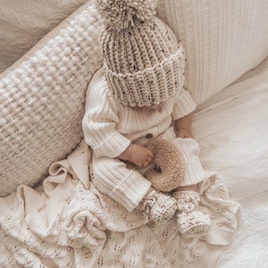May include: A baby wearing a cream-colored knit hat with a large pom-pom, matching sweater, pants, and booties. The baby is lying on a cream-colored blanket and pillow. A small stuffed toy is in the baby's hand.