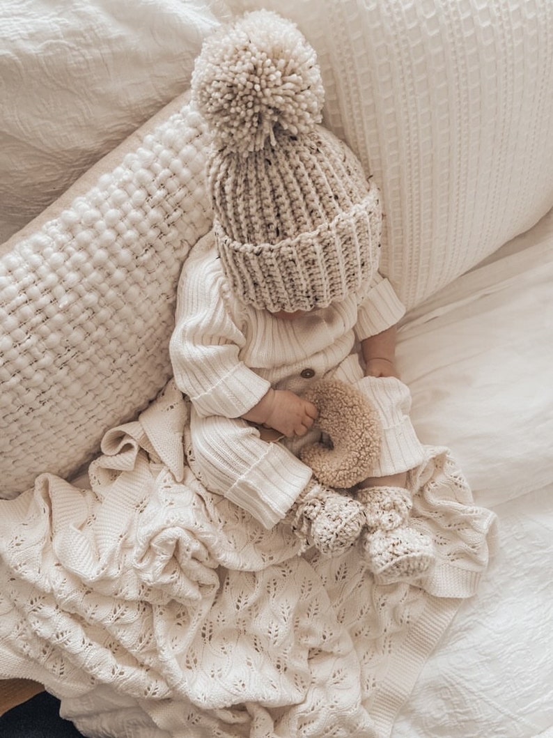 May include: A baby wearing a cream-colored knit hat with a large pom-pom, matching sweater, pants, and booties. The baby is lying on a cream-colored blanket and pillow. A small stuffed toy is in the baby's hand.