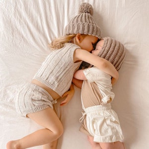 May include: Two children wearing matching beige knit outfits and hats with pom-poms, lying on a white sheet. One child wears a ribbed knit top and shorts, while the other wears a short-sleeved shirt and shorts. The children are embracing.