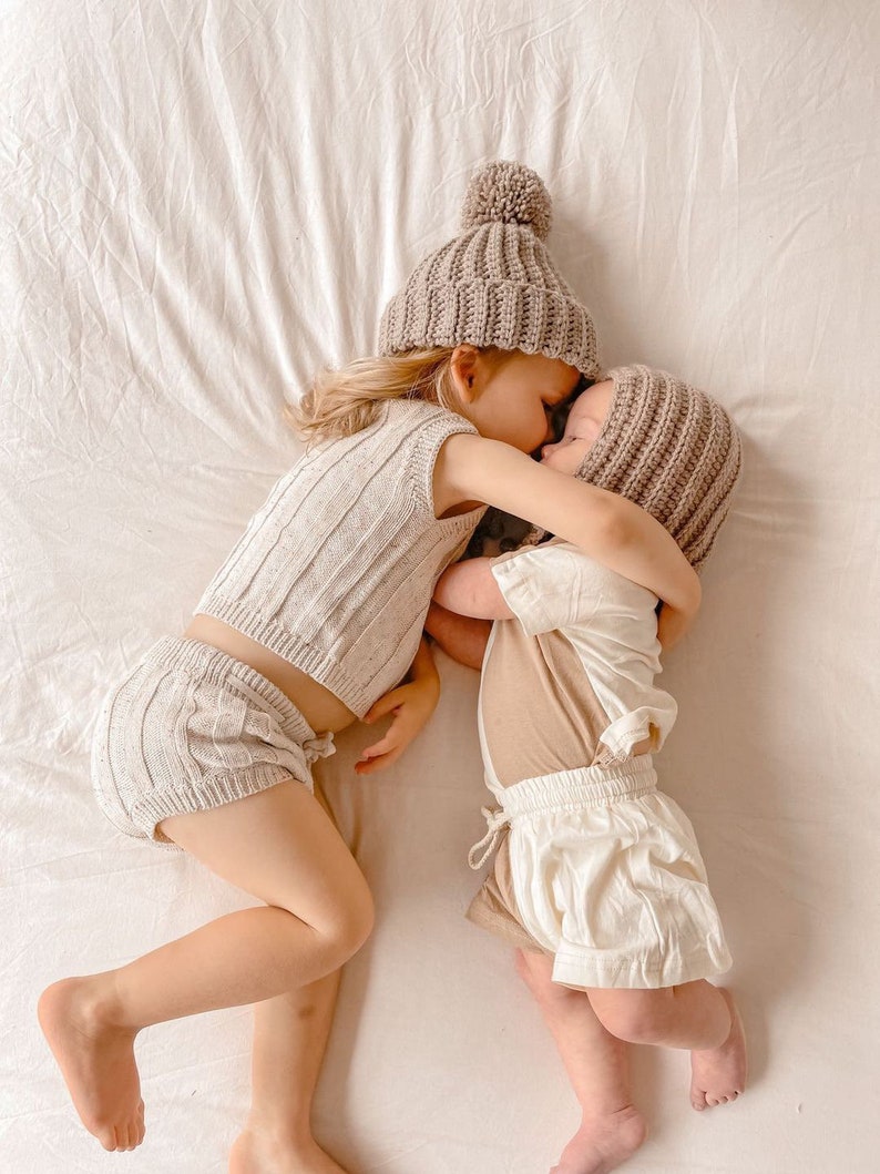 May include: Two children wearing matching beige knit outfits and hats with pom-poms, lying on a white sheet. One child wears a ribbed knit top and shorts, while the other wears a short-sleeved shirt and shorts. The children are embracing.