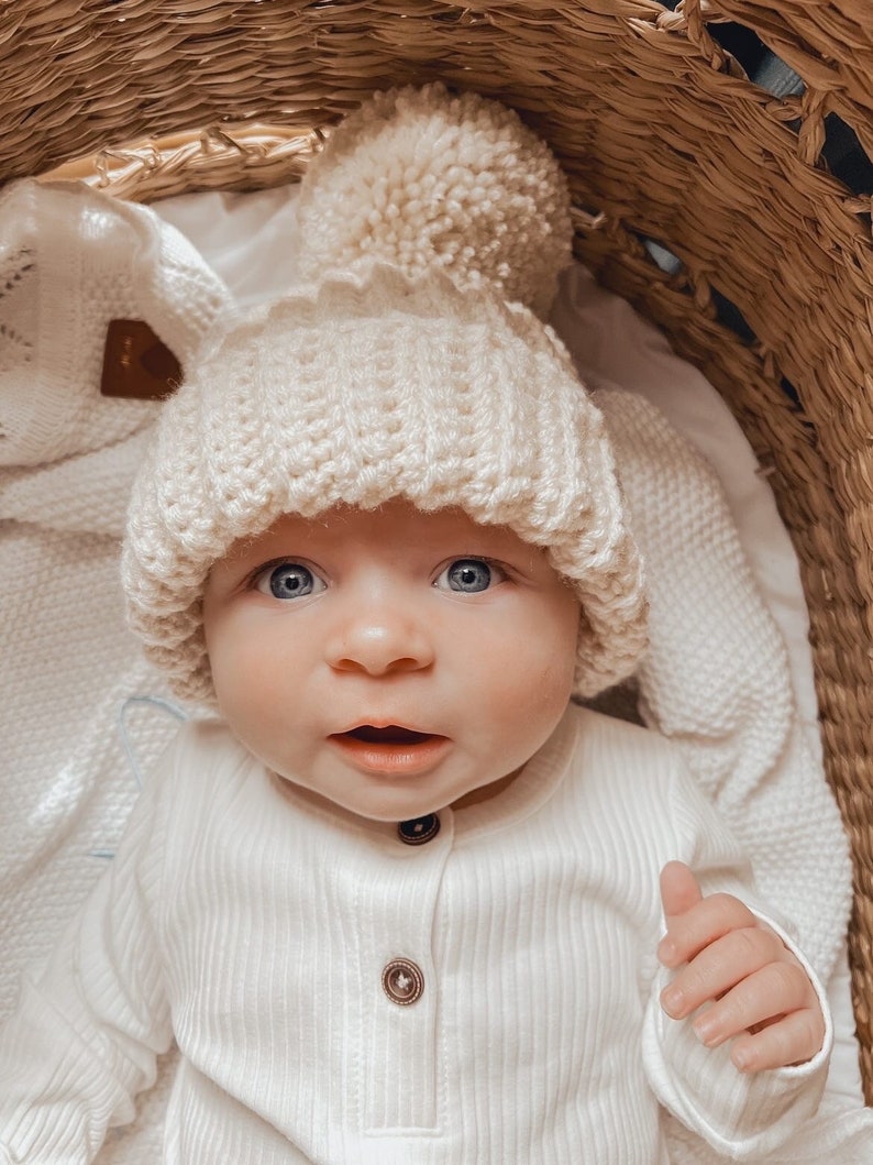 May include: A baby wearing a cream-colored knit hat with a large pom-pom and a cream-colored ribbed onesie with wooden buttons. The baby is in a woven basket, with a white blanket.