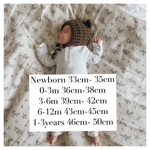 May include: A newborn baby wearing a brown crocheted bear hat and a white ribbed onesie, lying on a floral patterned blanket. The hat has small bear ears. Text on the image indicates hat sizes from newborn to 1-3 years.