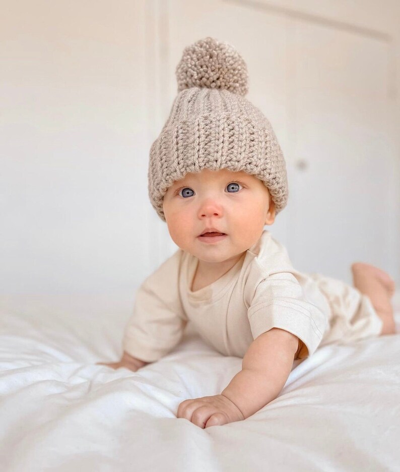 May include: A baby wearing a beige knit beanie with a large pom-pom and a beige onesie. The baby is lying on a white bedspread, looking directly at the camera.