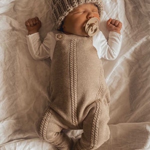 May include: A newborn baby wearing a beige knitted beanie with a pom-pom, a matching knitted romper, and a pacifier. The baby is lying on a white sheet.