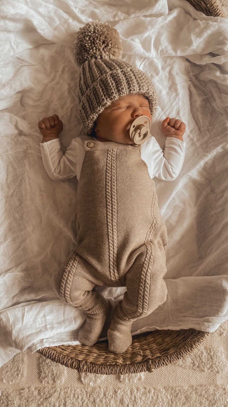 May include: A newborn baby wearing a beige knitted beanie with a pom-pom, a matching knitted romper, and a pacifier. The baby is lying on a white sheet.