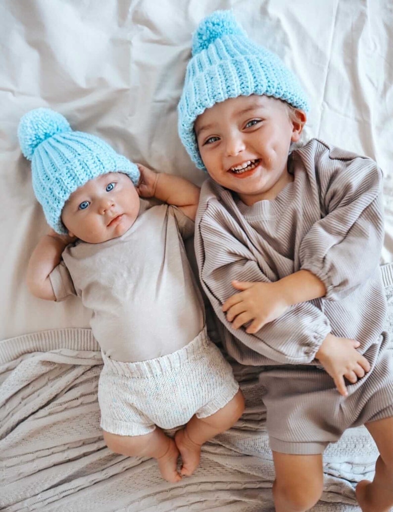 May include: Two babies wearing matching light blue knit hats with pom-poms. One baby is wearing a beige onesie and white knit shorts. The other baby is wearing a beige ribbed long-sleeve shirt and shorts set. Both babies are smiling.