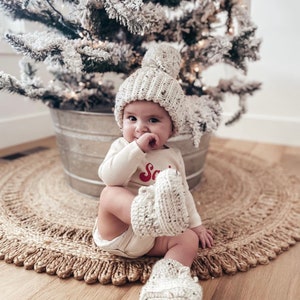 May include: A baby wearing a cream-colored knit hat with a pom-pom, matching booties, and a long-sleeved onesie with the word "Santa" in red. The baby is sitting on a woven rug near a snowy Christmas tree.