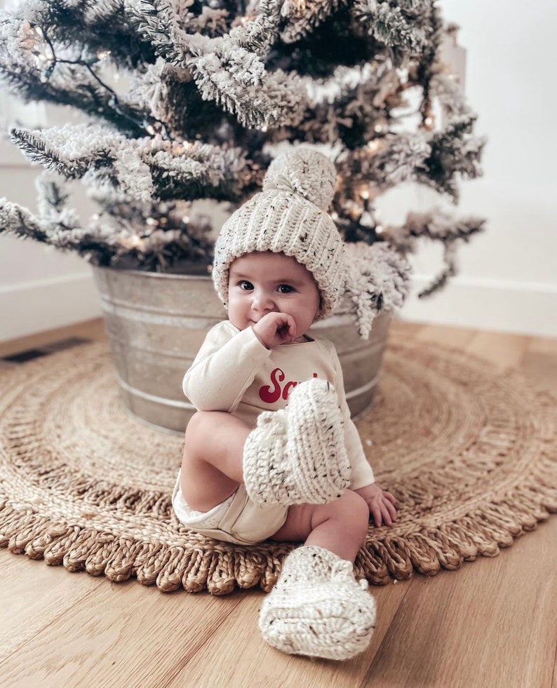 May include: A baby wearing a cream-colored knit hat with a pom-pom, matching booties, and a long-sleeved onesie with the word "Santa" in red. The baby is sitting on a woven rug near a snowy Christmas tree.