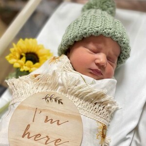 May include: A newborn baby wrapped in a sunflower-patterned blanket and wearing a green knitted hat with a pom-pom. A wooden sign with the words "I'm here" is in the foreground. The baby is lying on a white sheet, with a sunflower in the background.
