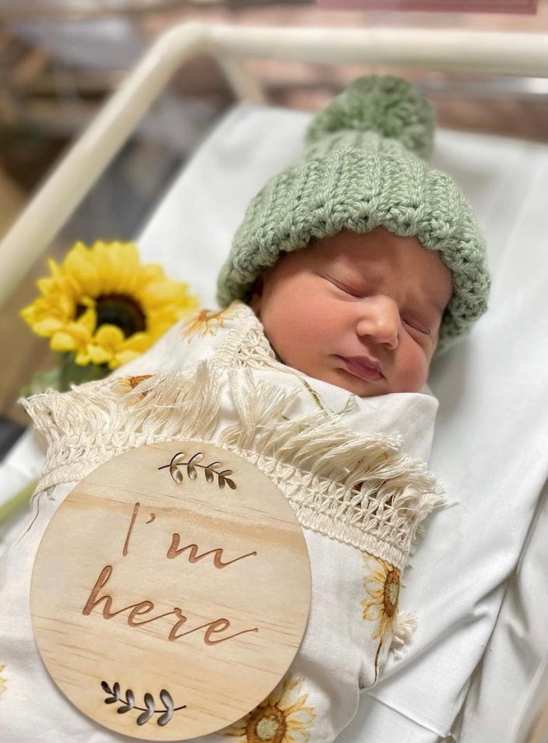 May include: A newborn baby wrapped in a sunflower-patterned blanket and wearing a green knitted hat with a pom-pom. A wooden sign with the words "I'm here" is in the foreground. The baby is lying on a white sheet, with a sunflower in the background.