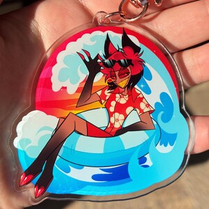 May include: A clear acrylic keychain featuring a cartoon character with red hair and black ears wearing sunglasses and a red floral shirt. The character sits in a blue and white striped inner tube amidst blue waves against a sunset background.