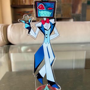 Puede incluir: Una figura de acr&iacute;lico transparente de un personaje de dibujos animados con una cabeza de pantalla de televisi&oacute;n, que lleva un traje azul y blanco con una pajarita roja. La figura est&aacute; de pie sobre una base con las letras "A" y "V" en un dise&ntilde;o rojo y azul. La pantalla muestra un ojo rojo y una boca dentada.