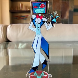 Puede incluir: Una figura de acr&iacute;lico transparente de un personaje de dibujos animados con una cabeza de pantalla de televisi&oacute;n, que lleva un traje azul y blanco con una pajarita roja. La figura est&aacute; sobre una base transparente con un logotipo rojo y turquesa.