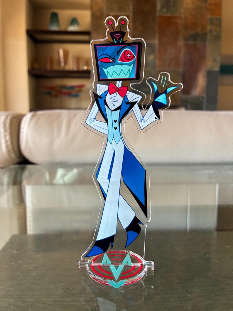 Puede incluir: Una figura de acr&iacute;lico transparente de un personaje de dibujos animados con una cabeza de pantalla de televisi&oacute;n, que lleva un traje azul y blanco con una pajarita roja. La figura est&aacute; sobre una base transparente con un logotipo rojo y turquesa.