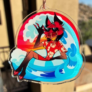 May include: A clear acrylic keychain featuring a cartoon character with red hair and black ears wearing sunglasses and a red floral shirt. The character sits in a blue and white striped inner tube surrounded by stylized blue waves and a sunset background.