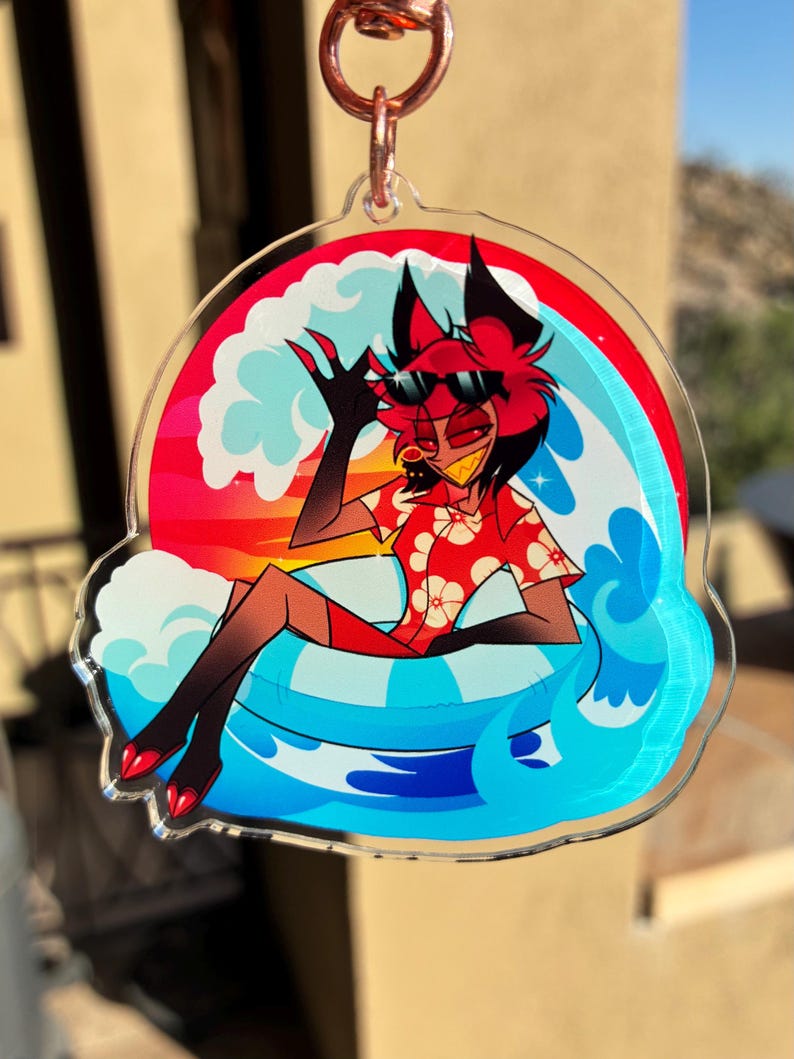May include: A clear acrylic keychain featuring a cartoon character with red hair and black ears wearing sunglasses and a red floral shirt. The character sits in a blue and white striped inner tube surrounded by stylized blue waves and a sunset background.