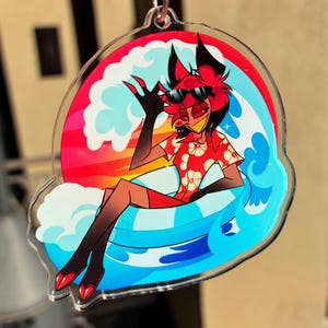 May include: A clear acrylic keychain featuring a cartoon character with red hair and black ears wearing black sunglasses and a red floral shirt. The character sits in a blue and white striped inner tube surrounded by stylized blue waves and a sunset.