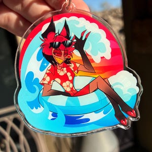 May include: A clear acrylic keychain featuring a cartoon character with red hair and black ears wearing black sunglasses and a red floral shirt. The character is lounging in a blue and white striped inner tube amidst stylized blue ocean waves.