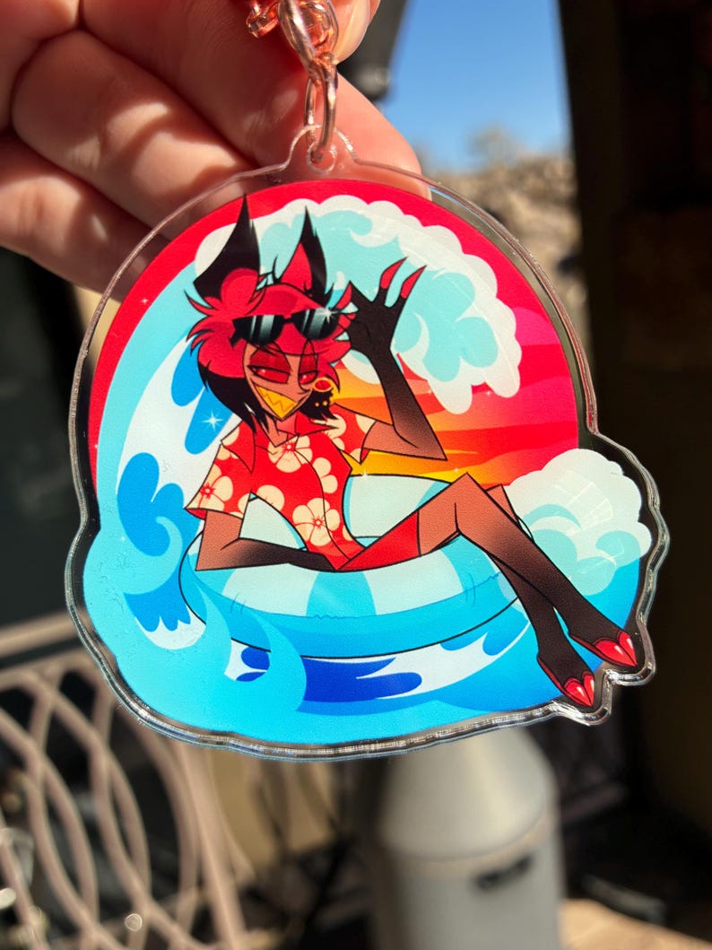 May include: A clear acrylic keychain featuring a cartoon character with red hair and black ears wearing black sunglasses and a red floral shirt. The character is lounging in a blue and white striped inner tube amidst stylized blue ocean waves.
