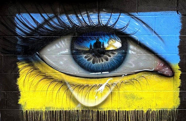 May include: A blue and yellow eye with a cityscape reflected in the pupil. The eye is painted on a brick wall and has a tear running down the cheek.