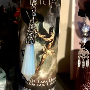 Könnte beinhalten: Eine weiße Kerze mit einem Schwarzweißbild einer geflügelten Figur mit dem Wort "Lucifer" auf dem Etikett. Eine silberne Kette mit einem blauen Kristallanhänger hängt an der Oberseite der Kerze.