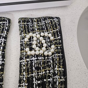 May include: Black and white tweed fingerless gloves with gold thread accents. The gloves feature a pearl embellishment in the shape of a double C logo. The gloves have a black leather trim.