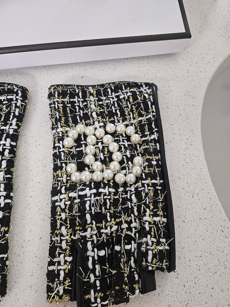 May include: Black and white tweed fingerless gloves with gold thread accents. The gloves feature a pearl embellishment in the shape of a double C logo. The gloves have a black leather trim.