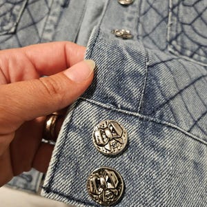 May include: A light blue denim jacket featuring a quilted diamond pattern across the fabric. The front placket is secured with round silver-toned metal buttons, each embossed with a detailed bow design and a small logo at the bottom center.