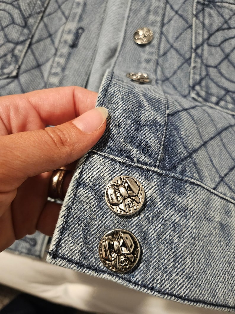 May include: A light blue denim jacket featuring a quilted diamond pattern across the fabric. The front placket is secured with round silver-toned metal buttons, each embossed with a detailed bow design and a small logo at the bottom center.