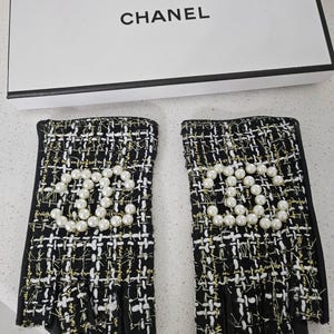May include: Black and white tweed fingerless gloves with gold accents and a pearl CC logo. The gloves are displayed with a white box that has the word "CHANEL" printed on it.