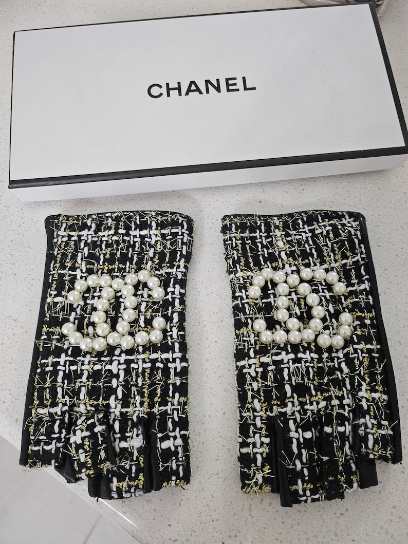May include: Black and white tweed fingerless gloves with gold accents and a pearl CC logo. The gloves are displayed with a white box that has the word "CHANEL" printed on it.
