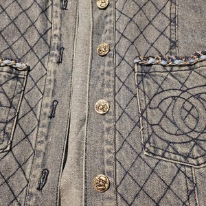May include: A light blue denim shirt featuring a diamond-stitched quilted pattern, silver-toned metallic buttons, and two chest pockets. Each pocket is accented with a braided trim of light blue and silver, and one pocket displays an embroidered logo.