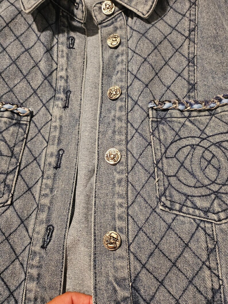 May include: A light blue denim shirt featuring a diamond-stitched quilted pattern, silver-toned metallic buttons, and two chest pockets. Each pocket is accented with a braided trim of light blue and silver, and one pocket displays an embroidered logo.